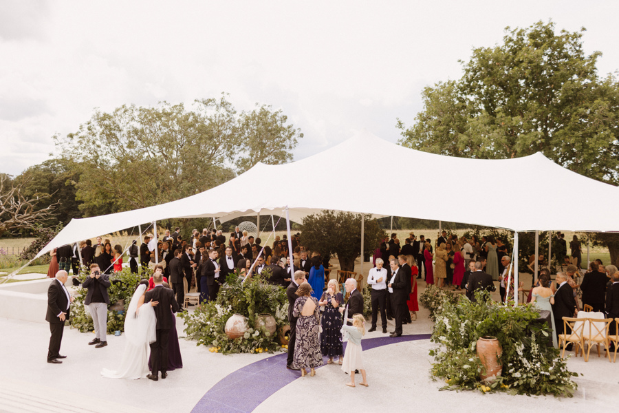 Wedding guests gathered under an open-sided stretch canopy in a garden