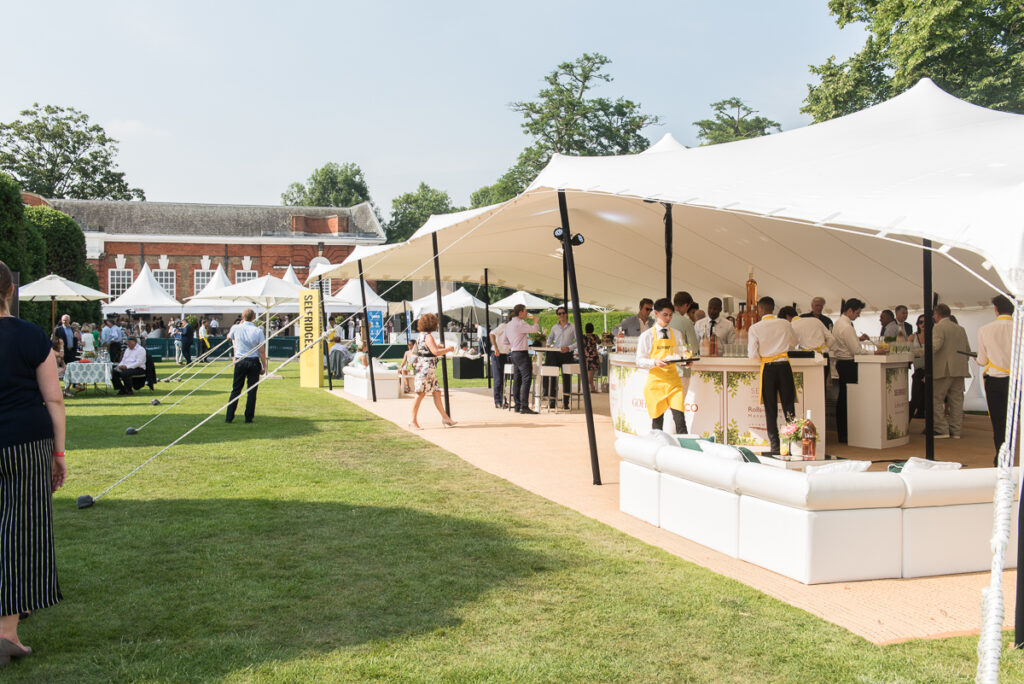 A white stretch tent sheltering champagne bars and white seating on a lawn beside a red-brick orangery