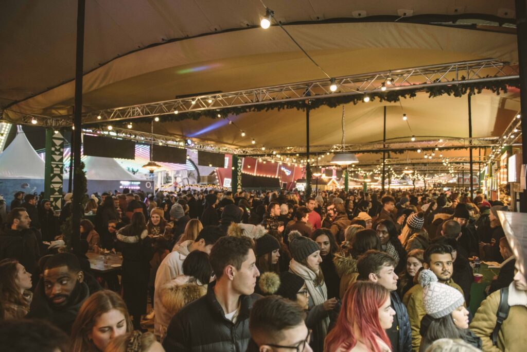 Crowded festival bar under a floodlit beige stretch tent at night