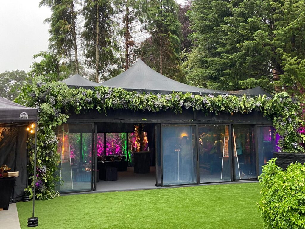 Glazed black pavilion dressed with foliage and lounge seating for a party